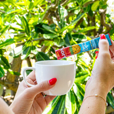 Hand pouring in a Lion Vanilla Macadamia Instant Coffee stick into a mug with greenery in the background.